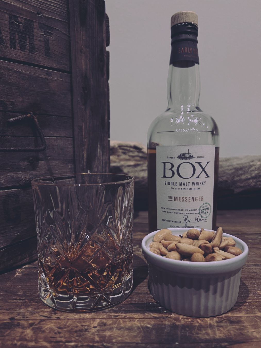 Ett glas med upphälld whisky och en liten skål jordnötter framför en whiskyflaska med texten ”BOX Single Malt Whisky The Messenger” stående på en skänk av trä, bredvid en större trälåda.