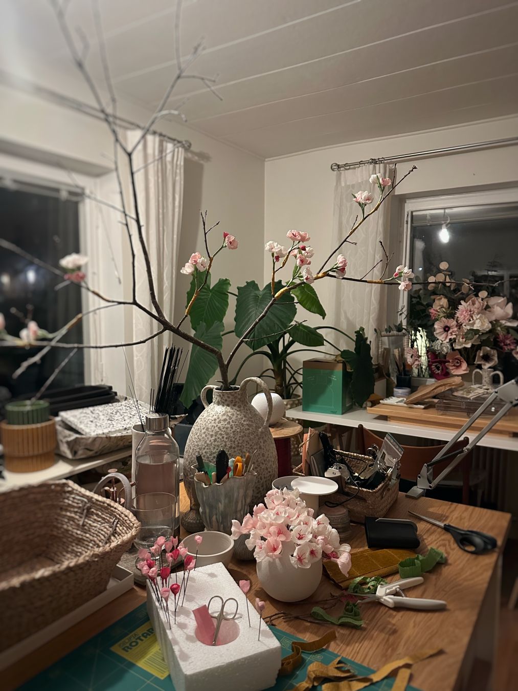 A large vase with a branch with some flowers on it and a small vase of paper flowers and buds in front. In the background a hobby room with various projects.