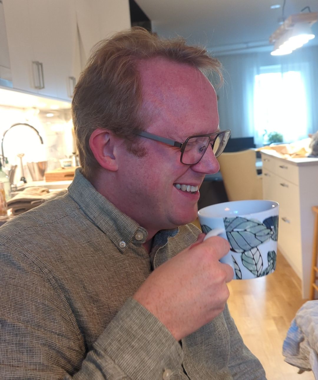 Foto i profil av en man som håller i en tekopp. Han ler stort mitt i ett skratt med ögonen hopknipna.