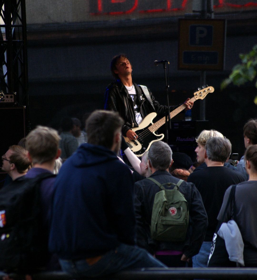 En ung man i vit ti-shirt och svart skinnjacka står bakom en mikrofon på en scen och spelar elbas. I förgrunden syns publik. Den unge mannen står med huvudet lätt tillbakalutat och blicken riktad ut mot publiken.