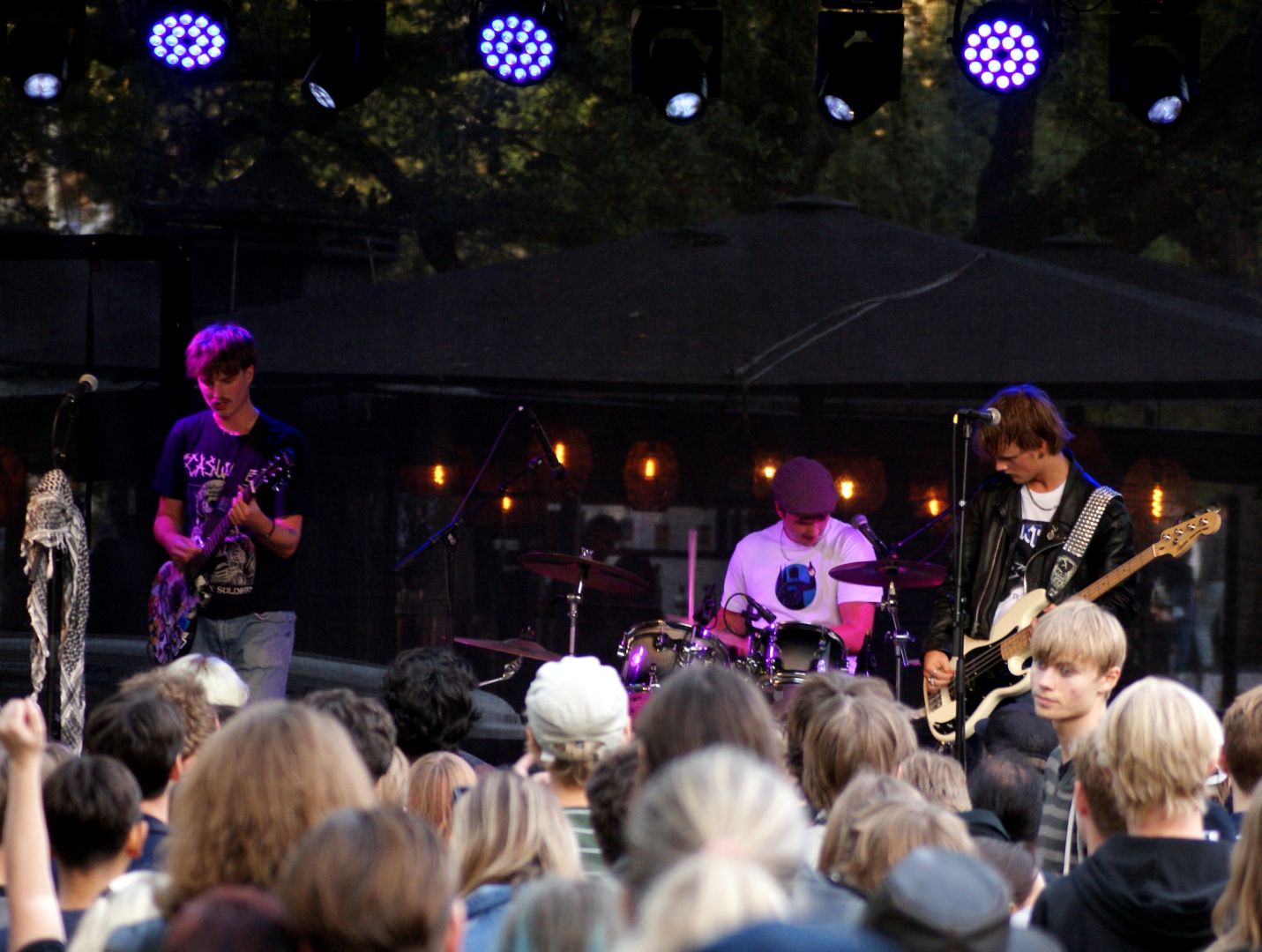 Samma band som i förra bilden, här fotade i vidvinkel så alla medlemmarna syns. Längst till vänster en ung man med kort hår och mustasch i svart t-shirt som spelar elgitarr bakom ett mikstativ med en palestinasjal på. I mitten längst bak sitter en trummis som har vit t-shirt och en gubbkeps. Längs till höger syns basisten från förra bilden.

I förgrunden syns huvuden på publiken.