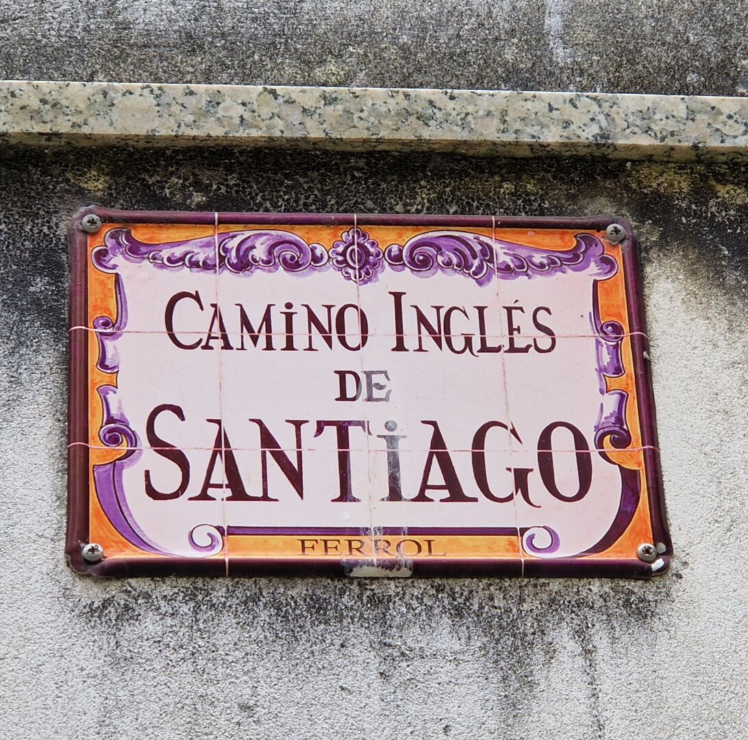 Blekt liten väggskylt i metall eller keramik med texten Camino Inglés de Santiago. Texten är inramad av lite krusiduller i orange och lila. Längst ned i det orange färgfältet står det Ferrol.