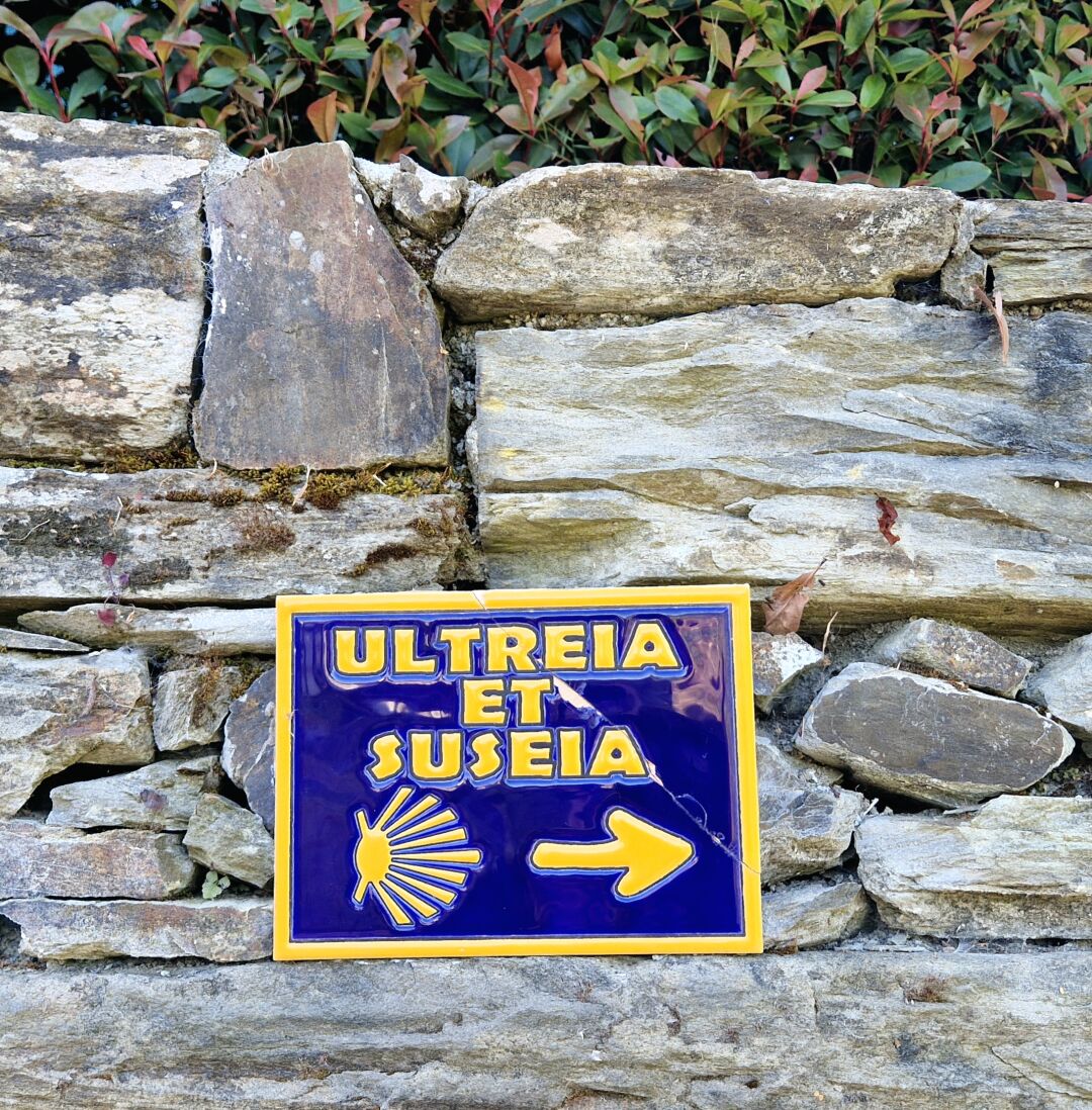 En liten keramikskylt i blått och gult med texten Ultreia et suseia  samt en bild av en pilgrimssnäcka och en pil. Uttrycket är en gammal hälsningsfras pilgrimer emellan och betyder Gå längre! Och gå högre!