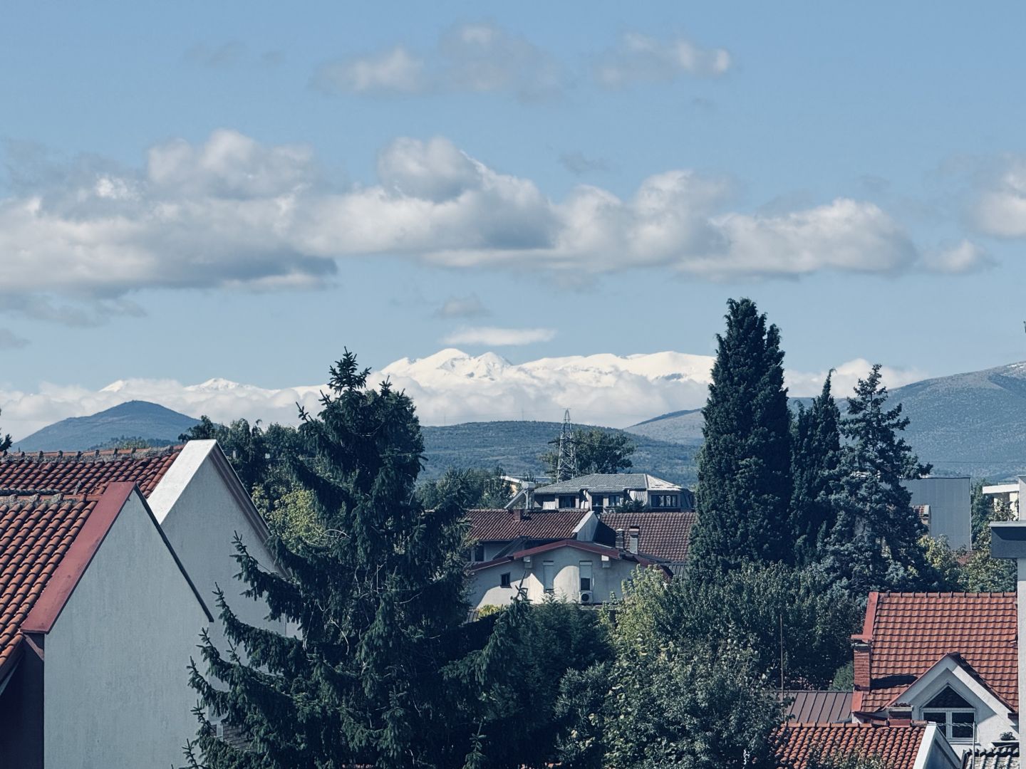 A landscape format photo of roof tops and urban fabric close to the camera. In the far distance one can see a snow covered mountain range. Above a blue sky with some light clouds. The sun is shining (not visible in picture)