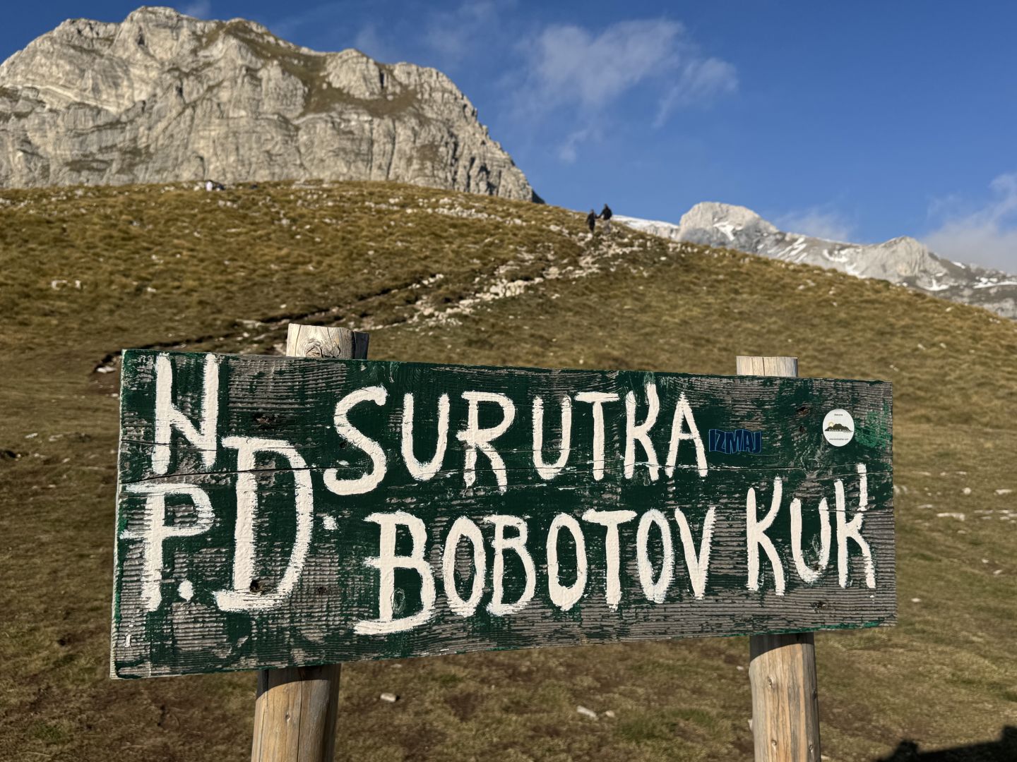 Picture of a sign in the Durmitor National Park. The sign says ”Surutka Bobotov Kuk”. Mountain visible in the background. Blue sky.
