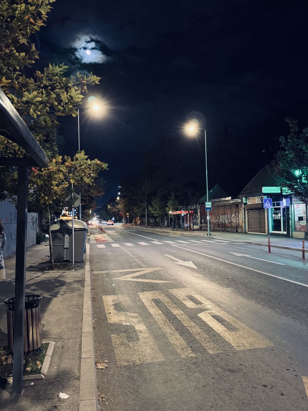 А street during night. A bus stop. “Bus” can be seen written in the picture upside down as the photographer is looking down the street towards where the bus should arrive from. Sky is dark. Streetlights are shining.