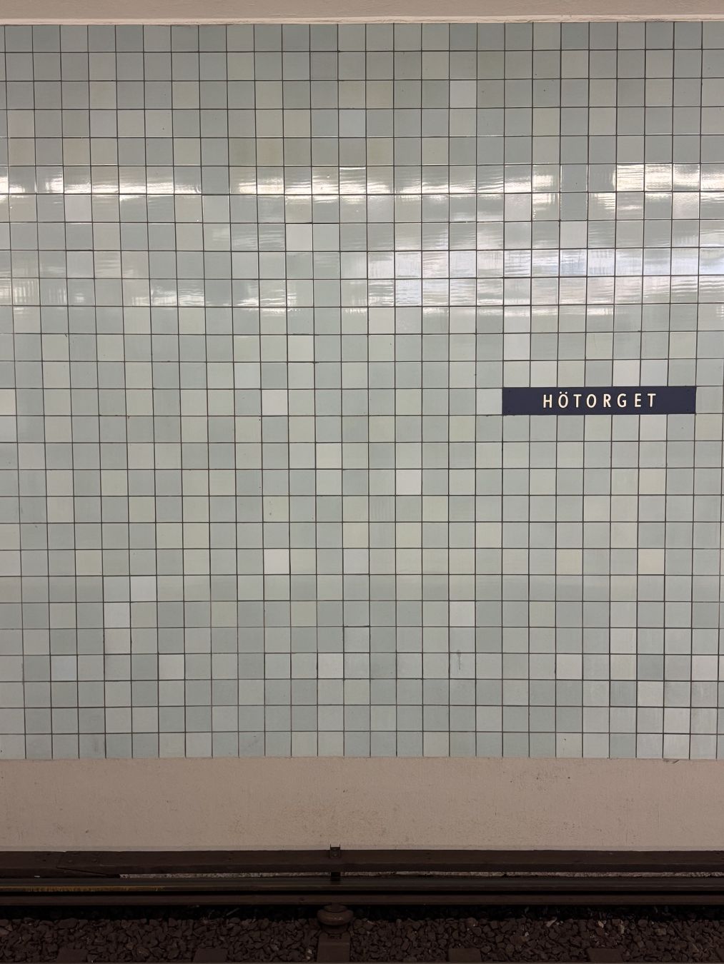 Photo of a wall in a metro station. The name of the metro station is visible on a dark blue sign with white letters saying ”Hötorget”. One of the more central stations in Stockholm. The wall is filled with tiles in a light bluish tone. There is a variation in level of blue. Tiles measure about 10x10 cm or 15x15 cm. Reflection of platform lighting can be seen in the upper part of the tile filled wall.
