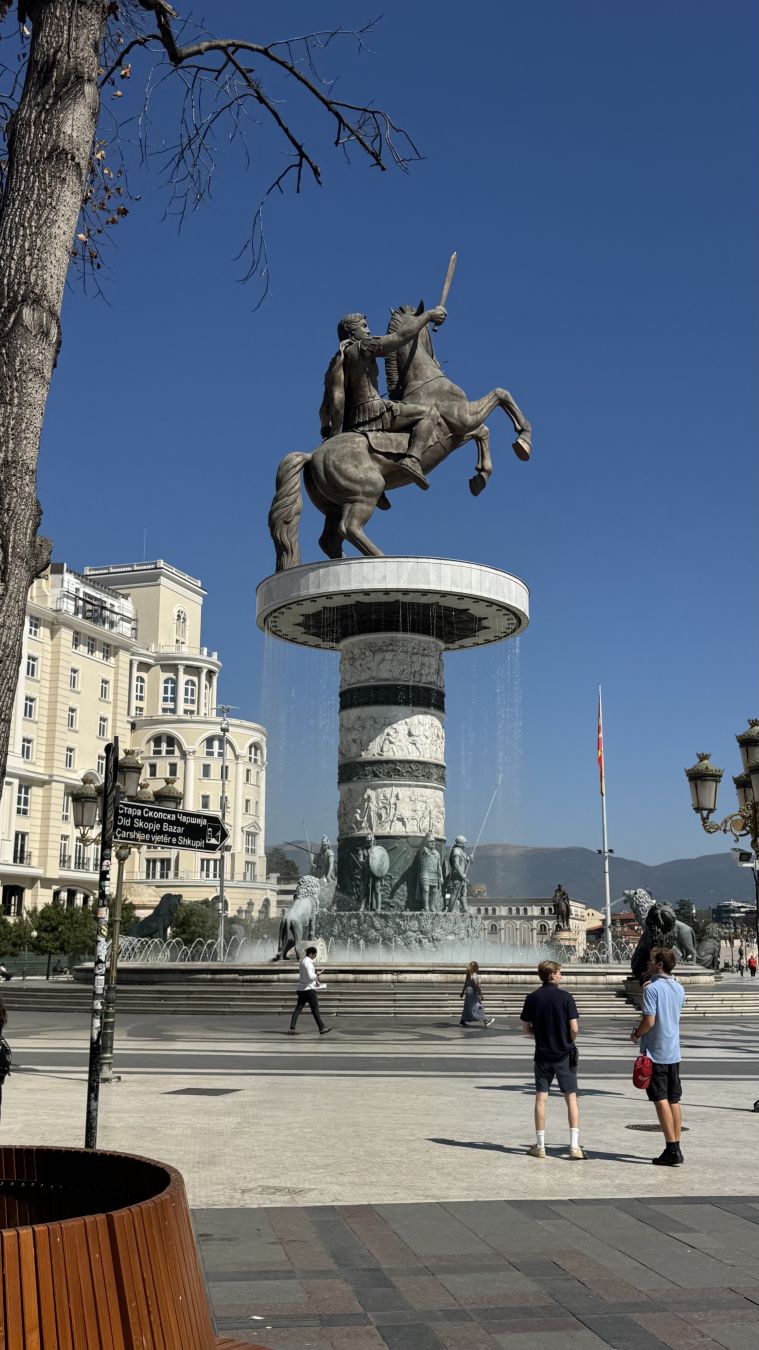 A statue is centered in the photo. A pillar holds a prancing horse with a male rider in its’ saddle. It’s Alexander The Great. Statue seems to be made out of bronze. 

Around the pillar is Macedonian soldiers from the time of Alexander’s life. A fountain surrounds the statue. 

A few people can be seen walking in the public space.