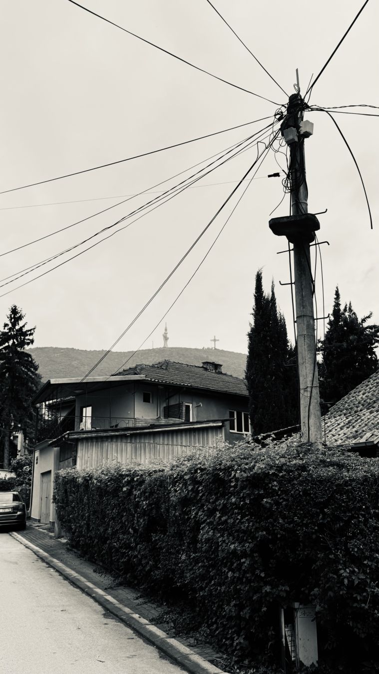 A black and white photo of an urban street. A pole for power lines is visible in the foreground. Some houses in the middle and in the far background one can see the Millennium Cross and new view tower at the top of Mount Vodno.