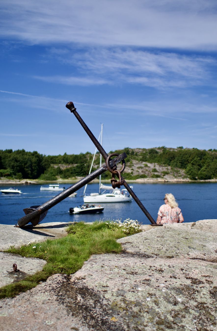 En kvinna sitter på en klippa vid sidan av ett stort ankare. Utanför är vatteninfarten till Grebbestad. På vattnet är en del båtar.